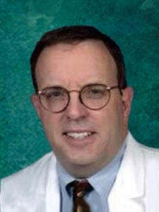 A man with short dark hair and glasses is wearing a white lab coat over a light brown tie and blue shirt. The background is a textured teal color.