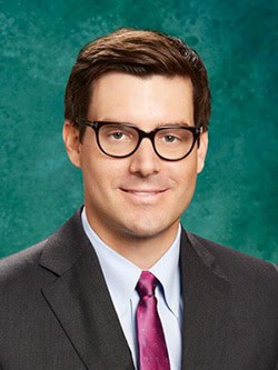 A man with short brown hair, wearing black-framed glasses, a dark suit, a light blue dress shirt, and a burgundy tie is smiling against a greenish-blue textured background.