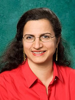 A woman with long, dark hair is smiling. She is wearing glasses, gold earrings, and a red collared shirt against a green background.