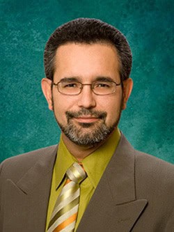 A man with short dark hair, a trimmed beard, and glasses is wearing a brown suit jacket, a mustard-yellow shirt, and a striped tie. He is posing against a teal, textured background.