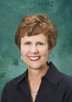 A woman with short, curly brown hair and a bright smile is wearing a dark collared shirt and dangly earrings. The background is a textured green color.