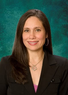 A woman with long, straight brown hair is smiling at the camera. She is wearing a black blazer, a pink top underneath, hoop earrings, and a gold necklace with a flower pendant. The background is a textured teal color.