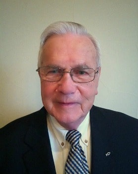 A senior man with white hair and glasses is wearing a dark suit, a light-colored dress shirt, and a striped tie. He is smiling slightly and looking directly at the camera against a plain background.