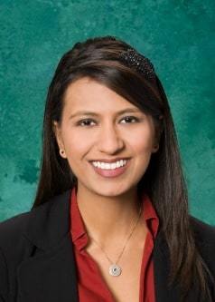 A woman with long brown hair, wearing a black blazer over a red top, smiles at the camera. She has a black hair accessory and a necklace with a circular pendant. The background is a textured teal color.