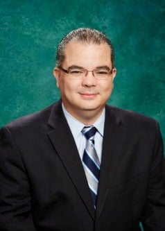 A man with short dark hair and glasses is wearing a dark suit, light blue shirt, and a blue striped tie. He is smiling slightly and posed against a green background.