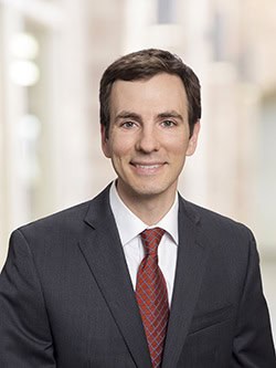 A man in a grey suit, white shirt, and red tie is smiling in a professional portrait. The background is blurred, featuring neutral tones and suggesting an office or indoor setting.
