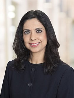 A woman with long, dark hair is smiling and facing the camera. She is wearing a black jacket with buttons on the front. The background is blurred and appears to be an indoor setting with neutral colors.