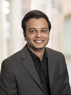 A man wearing a dark suit jacket and black shirt smiles at the camera. He has short, dark hair and is standing in front of a blurred indoor background.