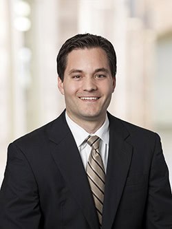 A man wearing a black suit, white shirt, and beige striped tie is smiling. He has short, dark hair and is posed in front of a blurred background with light, neutral tones.