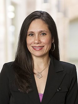 A woman with long, straight dark hair smiles at the camera. She is wearing a black blazer over a purple top, hoop earrings, and a floral pendant necklace. The background is softly blurred, suggesting an indoor setting with light-colored walls.
