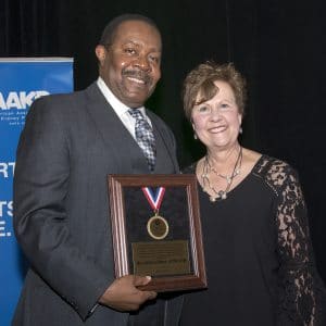 Two people pose for a photo in front of a dark background. The man on the left is holding a wooden plaque with a medal and smiling, wearing a gray suit and tie. The woman on the right is also smiling, wearing a black dress with lace sleeves. A blue banner is partially visible behind them.