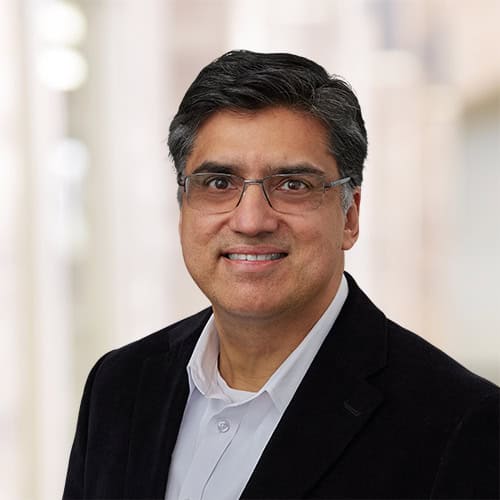 Syed-Ali Asghar, MD, with short dark hair and glasses, wearing a white shirt and black jacket, smiles at the camera. The background is softly blurred, featuring neutral tones.