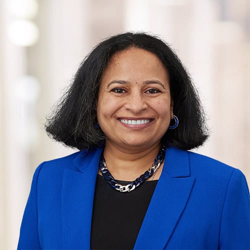 Dr. Archana Rao, MD, with medium-length black hair, smiles in a bright blue blazer, black top, and chunky black-and-silver necklace, standing before a blurred, light-colored background.