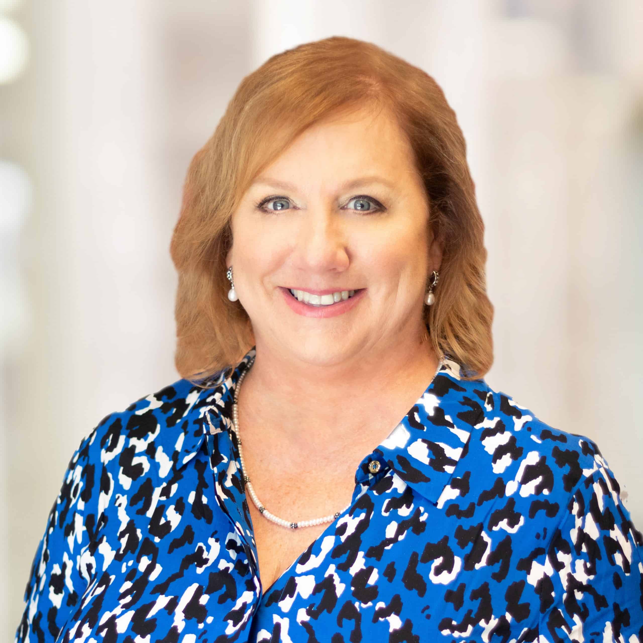 Dr. Sandra Lauriat with shoulder-length light brown hair smiles at the camera. She is wearing a blue and white patterned blouse and a necklace. The background is blurred, giving a soft and warm tone to the image.