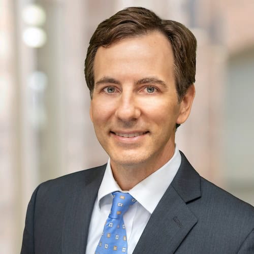 Dr. Richard C. Fuquay, MD, FASN, with brown hair, is wearing a dark suit, light blue shirt, and patterned light blue tie while smiling at the camera. The softly blurred background suggests an indoor setting with a neutral or light-colored backdrop.