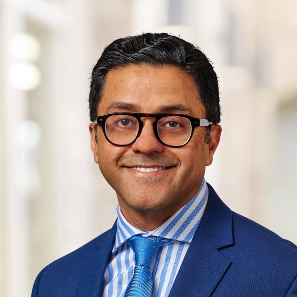 A professionally dressed man, resembling Salman Khan, with short black hair and glasses is smiling at the camera. He is wearing a blue suit, a striped blue shirt, and a blue tie. The background is blurred, suggesting an indoor setting, possibly an office.