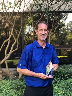 David Knop, in a blue polo shirt, stands outdoors in front of a building with large windows and trees. Smiling proudly, he holds the Arthur P. Pasquarella Leadership in Action Award. The background features vibrant greenery and well-trimmed bushes, adding to the celebratory atmosphere.