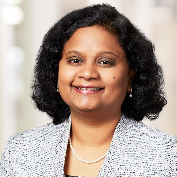 A woman with shoulder-length black curly hair, smiling, is wearing a light grey blazer over a dark top, with a pearl necklace and pearl earrings. The background is blurred, possibly depicting an indoor setting with bright lighting. This is Dr. Jayasree Grandhi, MD.
