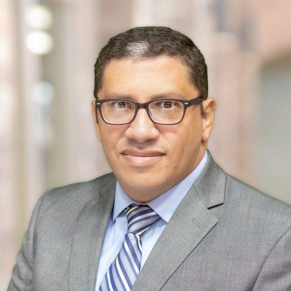 Dr. Wael Hanna, MD, a man with short dark hair and glasses, is wearing a grey suit with a light blue shirt and a striped blue and white tie. He is standing against a blurred indoor background, looking towards the camera with a neutral expression.