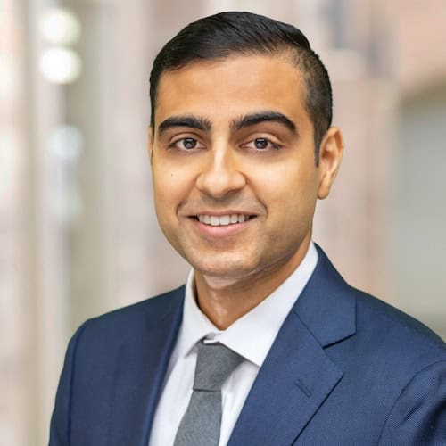 Vinay Acharya, DO, with short, dark hair and a light complexion, is smiling at the camera. He is wearing a dark blue suit, a white dress shirt, and a gray tie. The background is blurred, with light-colored walls and soft lighting.
