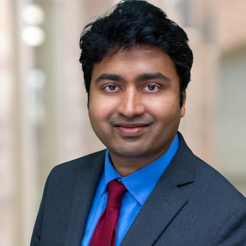 Suman Krishna Kotla, MD with short, dark hair is wearing a blue shirt, a red tie, and a dark suit jacket. They are looking into the camera with a slight smile. The background is blurred, giving the image a professional feel befitting an MD.