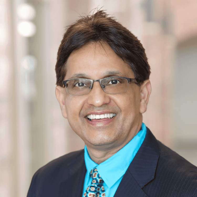 A man, identified as Arshad Asghar, MD, with short dark hair, glasses, and a big smile is wearing a black suit with a bright blue dress shirt and a colorful tie. The background is blurred and appears to be an indoor setting with soft lighting.