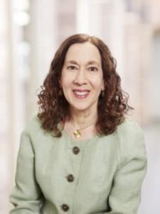 Dr. Levy wavy brown hair, wearing a light green blazer, smiles at the camera. She stands against a blurred background of light-colored walls and windows, embodying 50 years of excellence in kidney care.
