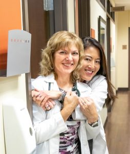 Two female healthcare professionals smile while standing in a clinic hallway. One, with curly blonde hair, wears a stethoscope and is being hugged from behind by her colleague with long, straight dark hair. Both are dressed in white lab coats and appear cheerful and friendly.