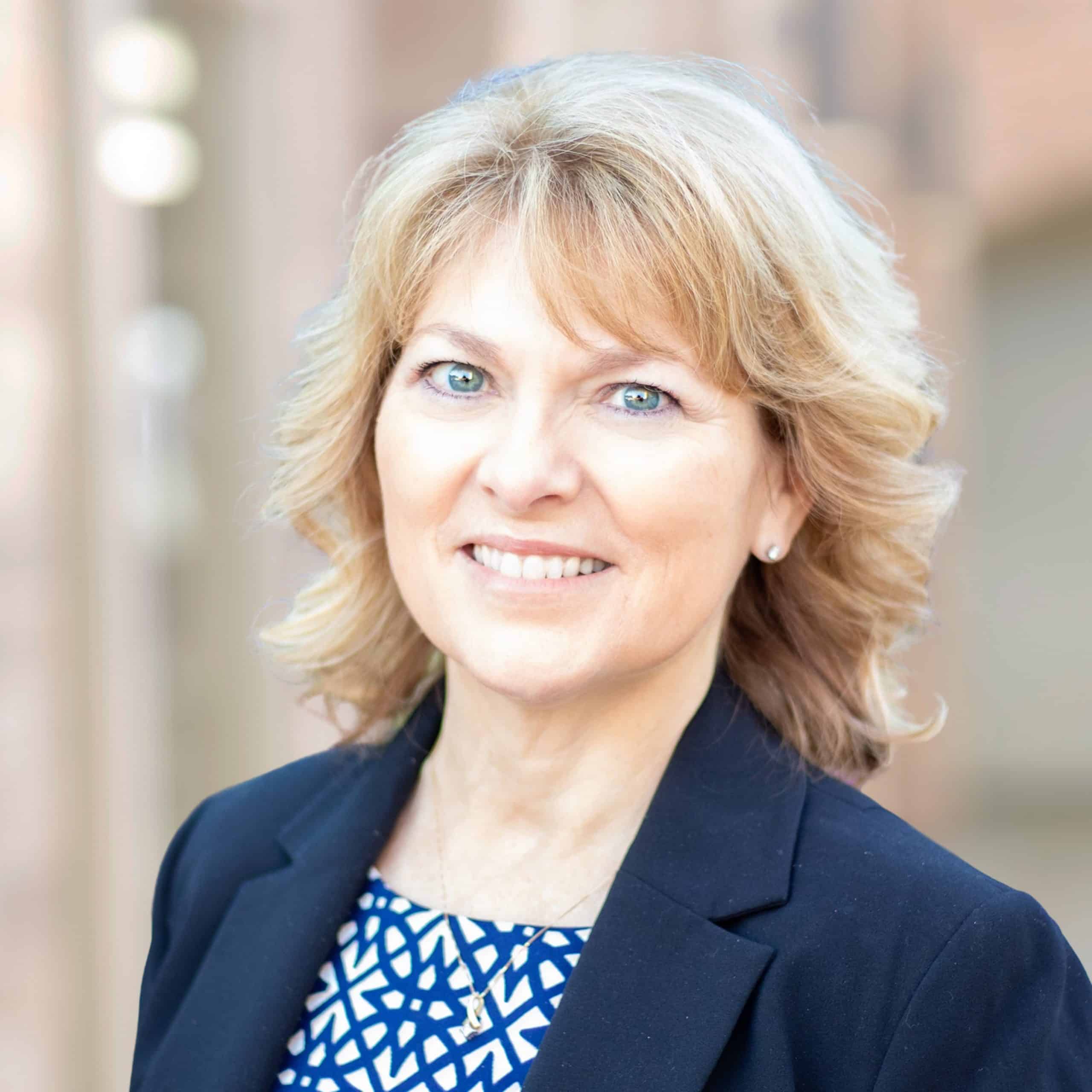 A smiling Teresa McLemore, PA-C, with shoulder-length blonde hair, is wearing a black blazer over a blue and white patterned blouse. The background is blurred, suggesting an outdoor setting with soft lighting.