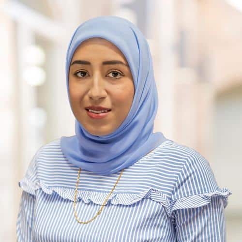 A woman, Wajeeha Sindhu, wearing a light blue hijab and a striped shirt with ruffles stands against a softly blurred background. She has a gold chain necklace and is smiling slightly.
