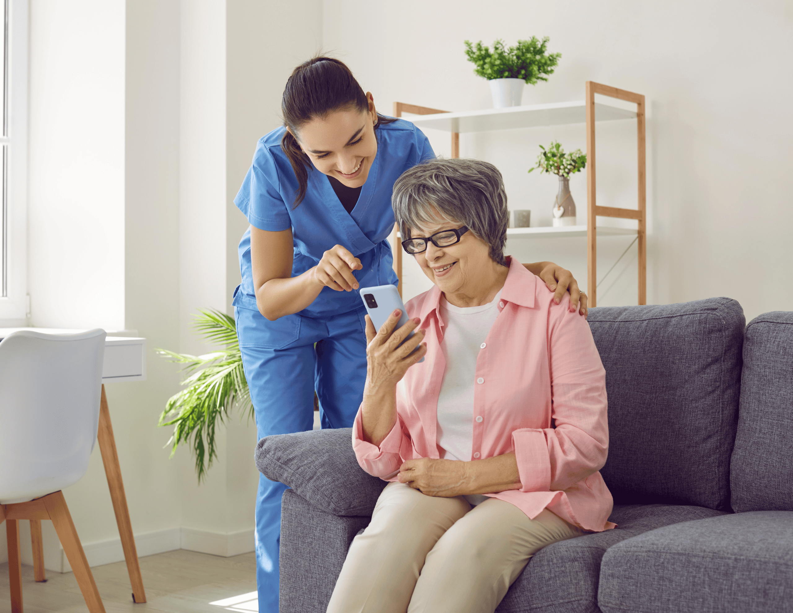 A healthcare worker in blue scrubs is standing next to an elderly woman in glasses and a pink shirt, who is sitting on a gray couch. They are both smiling while looking at a smartphone the elderly woman is holding. A white bookshelf is in the background.