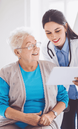 A smiling healthcare professional shows a tablet to an elderly woman in a blue shirt and beige cardigan, discussing diet and nutrition. They are in a well-lit room, indicating a positive, engaging interaction.
