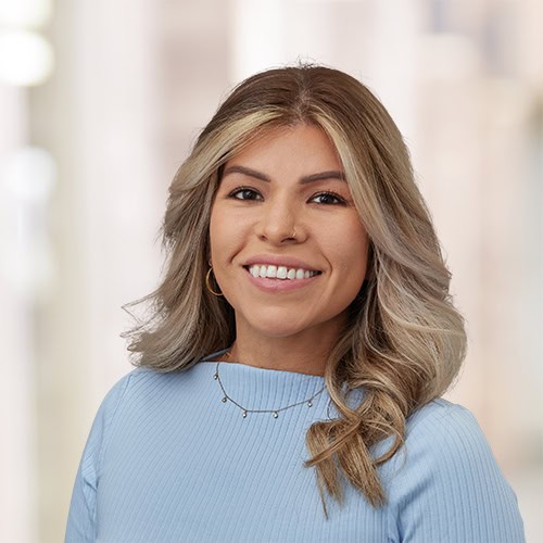 A woman with long, wavy blonde hair—Khrystal Padron, APRN, AGACNP-BC—wearing a light blue top and gold jewelry is smiling at the camera. The background is softly blurred, suggesting an indoor setting.