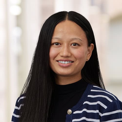A woman with long, straight dark hair smiles at the camera. She is wearing a navy blue and white striped sweater over a black top. The background is softly blurred with neutral tones.