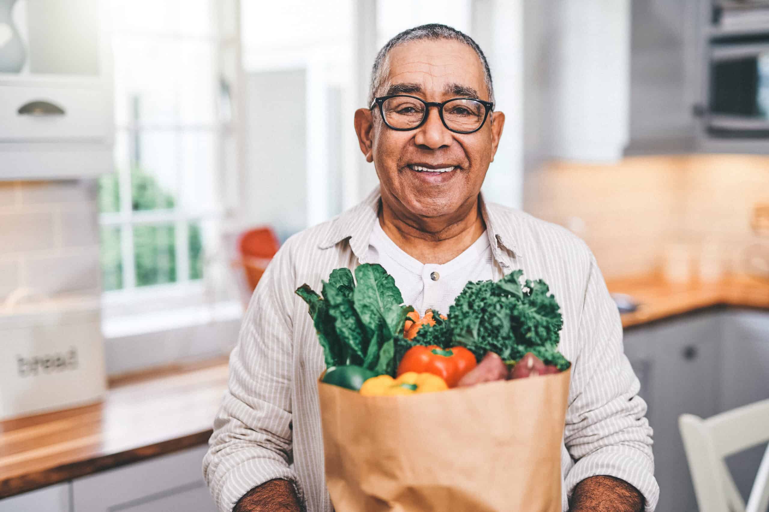 Smiling older man wearing glasses and a striped shirt stands in a kitchen, holding a paper bag filled with DASH Diet and Kidney Health-friendly vegetables like kale, bell peppers, and spinach. Bright natural light streams through the window behind him.