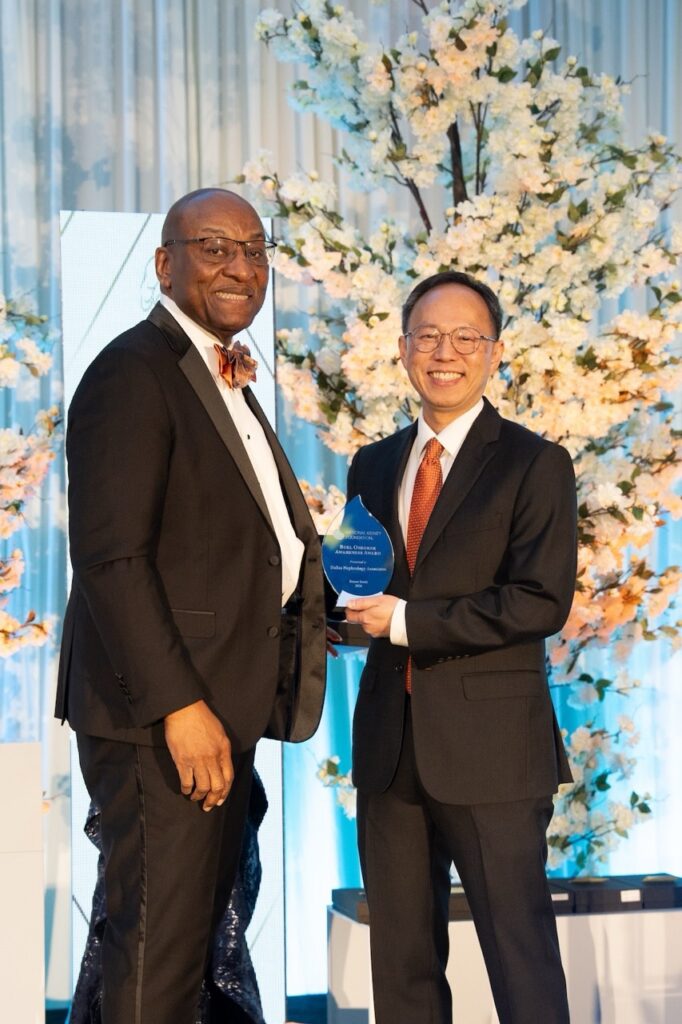 Two men in suits stand smiling at an event; one presents the Burl Osborne Award to the other. They are in front of a backdrop with decorative white flowers and blue lighting, both appearing happy and formal.