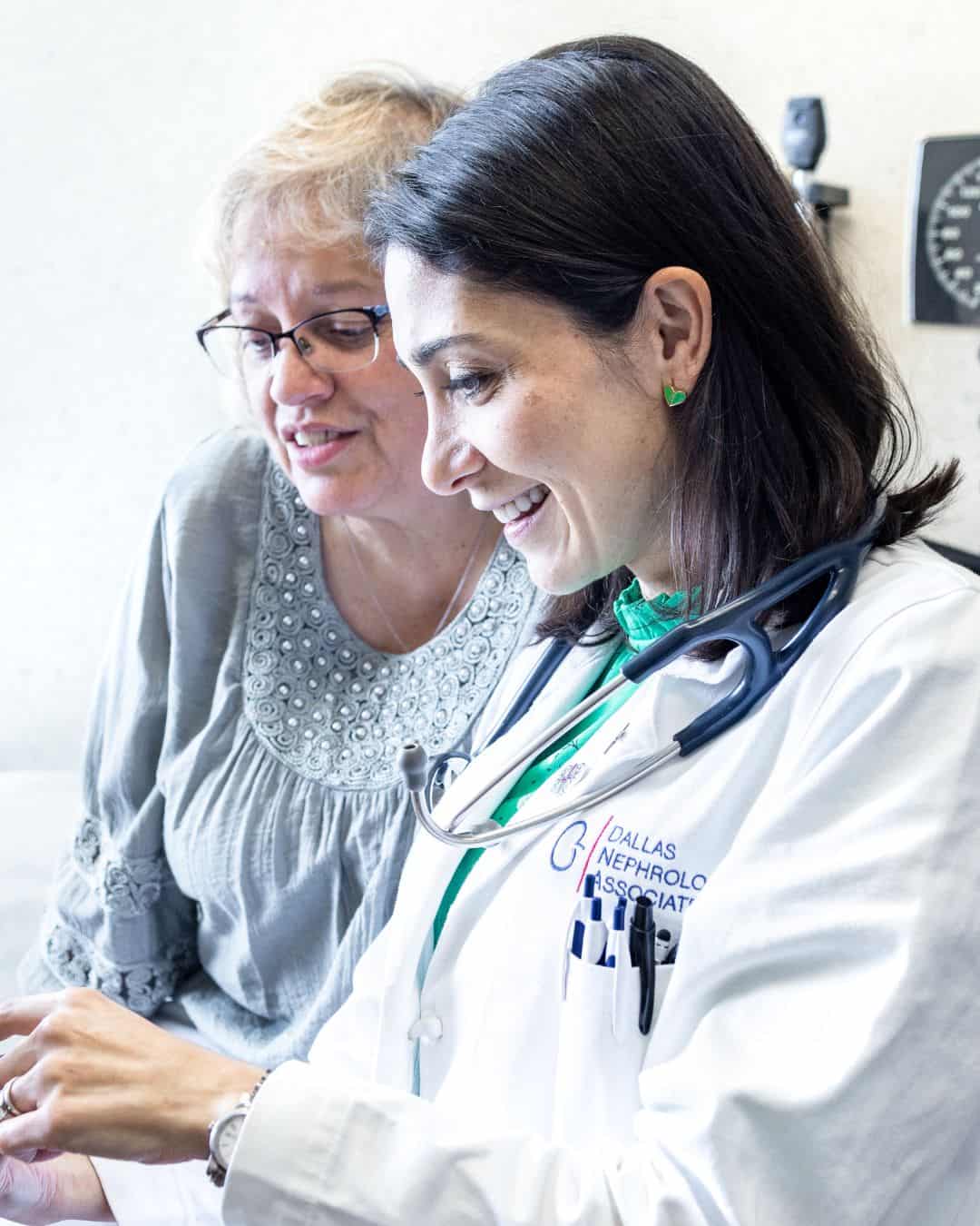 A doctor in a white coat with a stethoscope smiles while showing information to a patient, who is wearing glasses and a grey blouse. They appear to be discussing Chronic Kidney Disease results in a clinical setting.