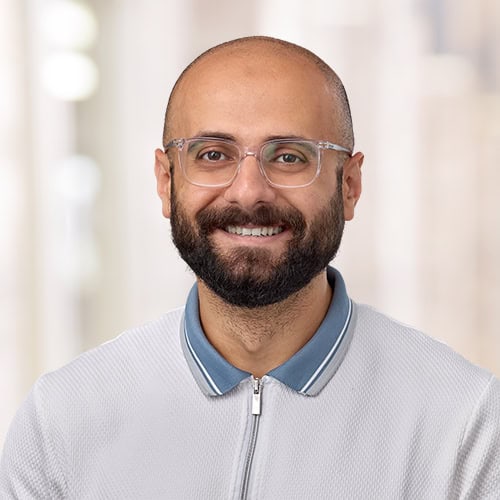 Smiling man with glasses, a beard, and a bald head, wearing a light gray zip-up shirt with a blue collar, standing against a blurred light background—identified as Mohammed Miqdad, MBBS.