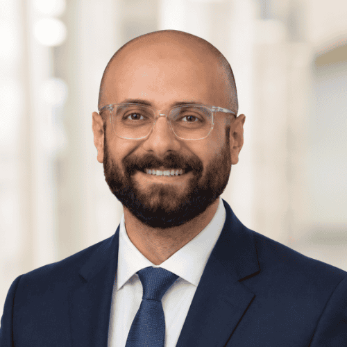 A man with a beard and glasses, wearing a navy suit, white shirt, and blue tie, smiles at the camera against a blurred indoor background—introducing Dr. Mohammed Miqdad, MBBS.