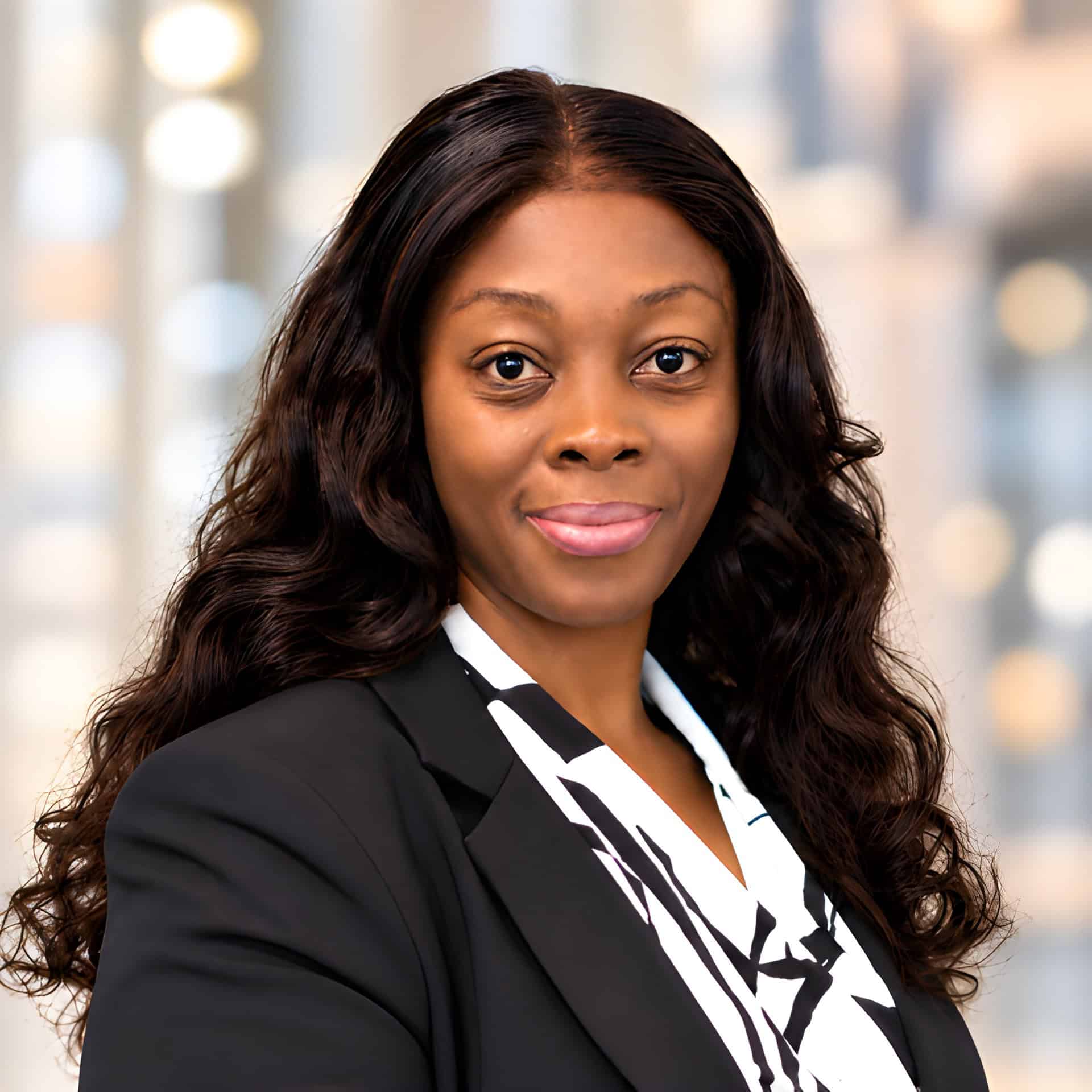 Belinda Mesape, APRN, AGACNP-BC, with long wavy hair and a soft smile, wears a black blazer over a patterned blouse. The warm lights in the blurred background add to the inviting atmosphere.
